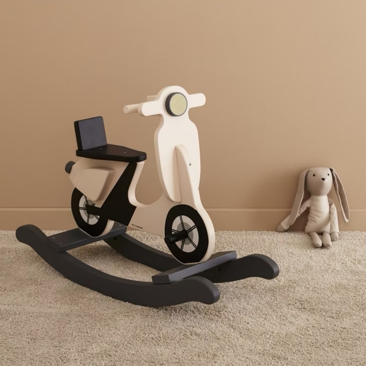 Wooden scooter-shaped rocking toy on a carpeted floor with a beige wall in the background.