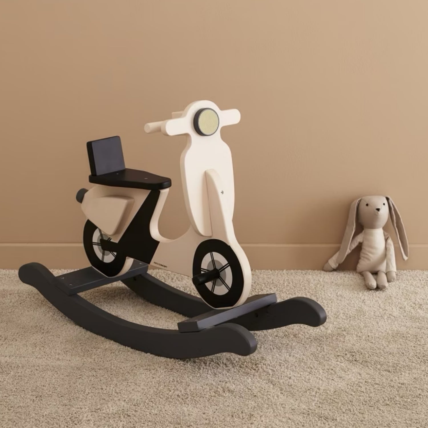 Wooden scooter-shaped rocking toy on a carpeted floor with a beige wall in the background.