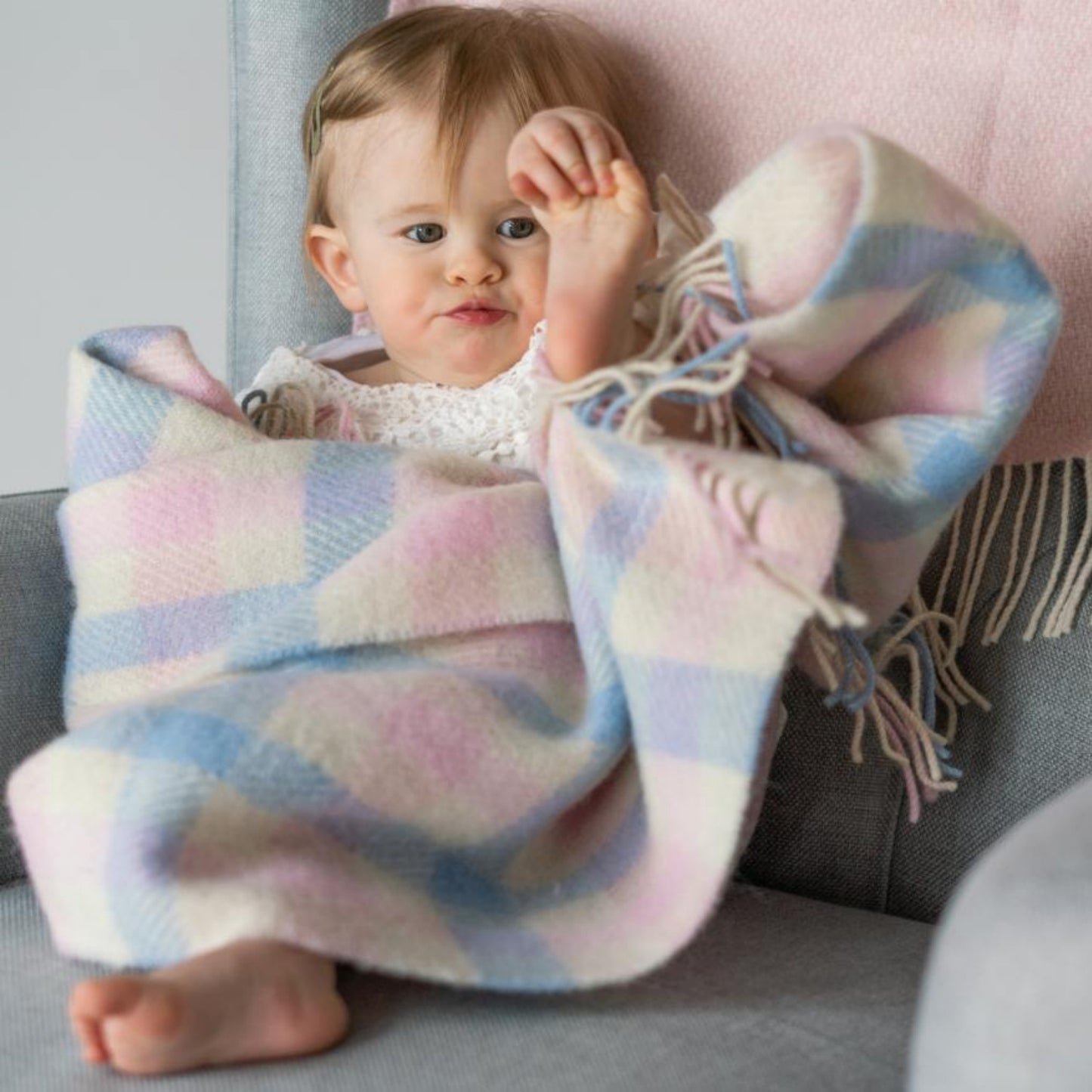 John Hanly Irish Wool Baby Blanket | White, Pink & Blue Border Block Check | 100% Pure Irish Wool Blanket | Lifestyle: Baby Playing on Sofa with Blanket | BeoVERDE Ireland